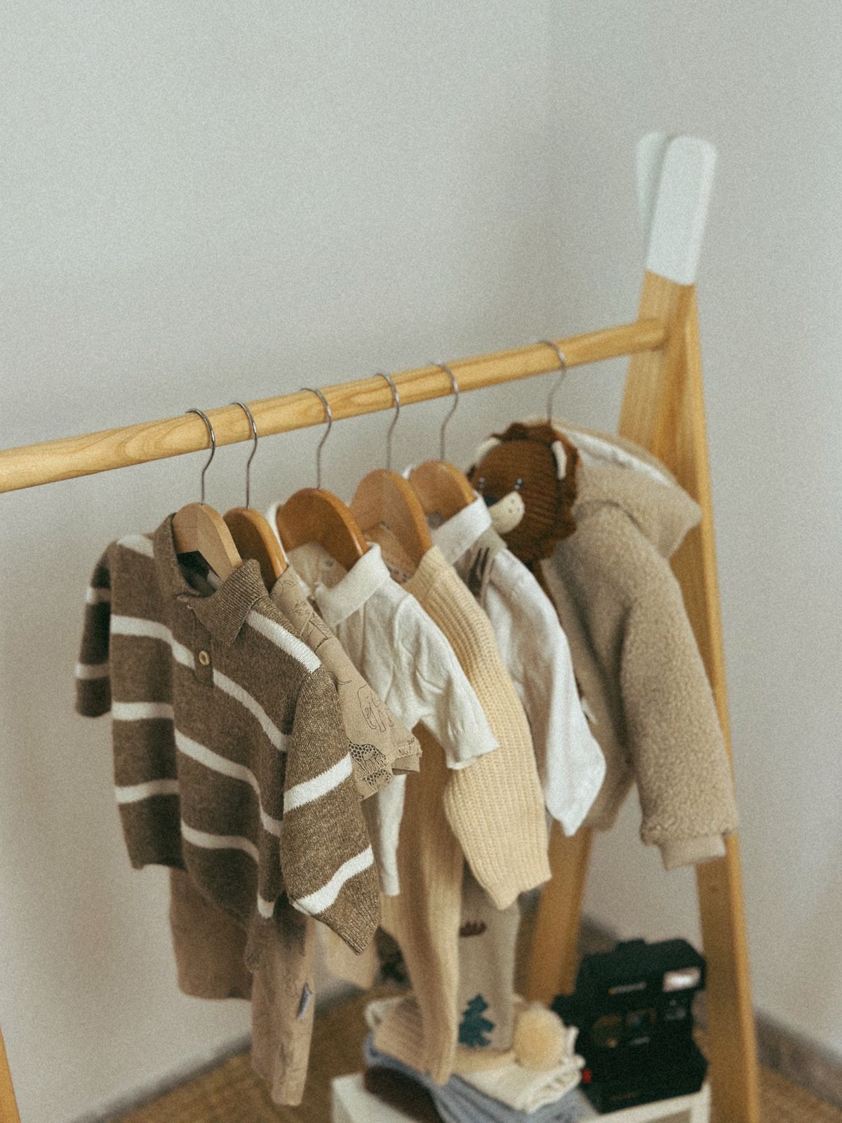Baby knits and cotton tops hanging on a timber rack against a pale wall.